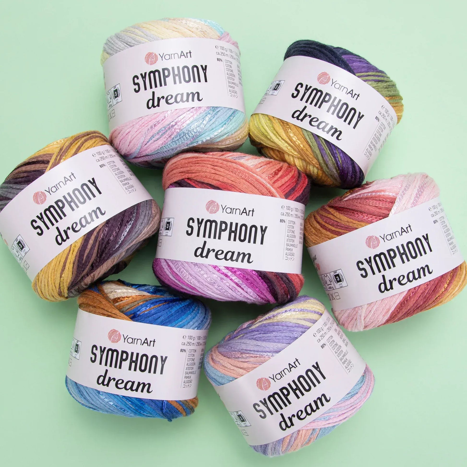 Conjunto de siete ovillos de hilado YarnArt Symphony Dream, mezcla de algodón y viscosa, en varios colores degradados vibrantes sobre fondo verde claro.