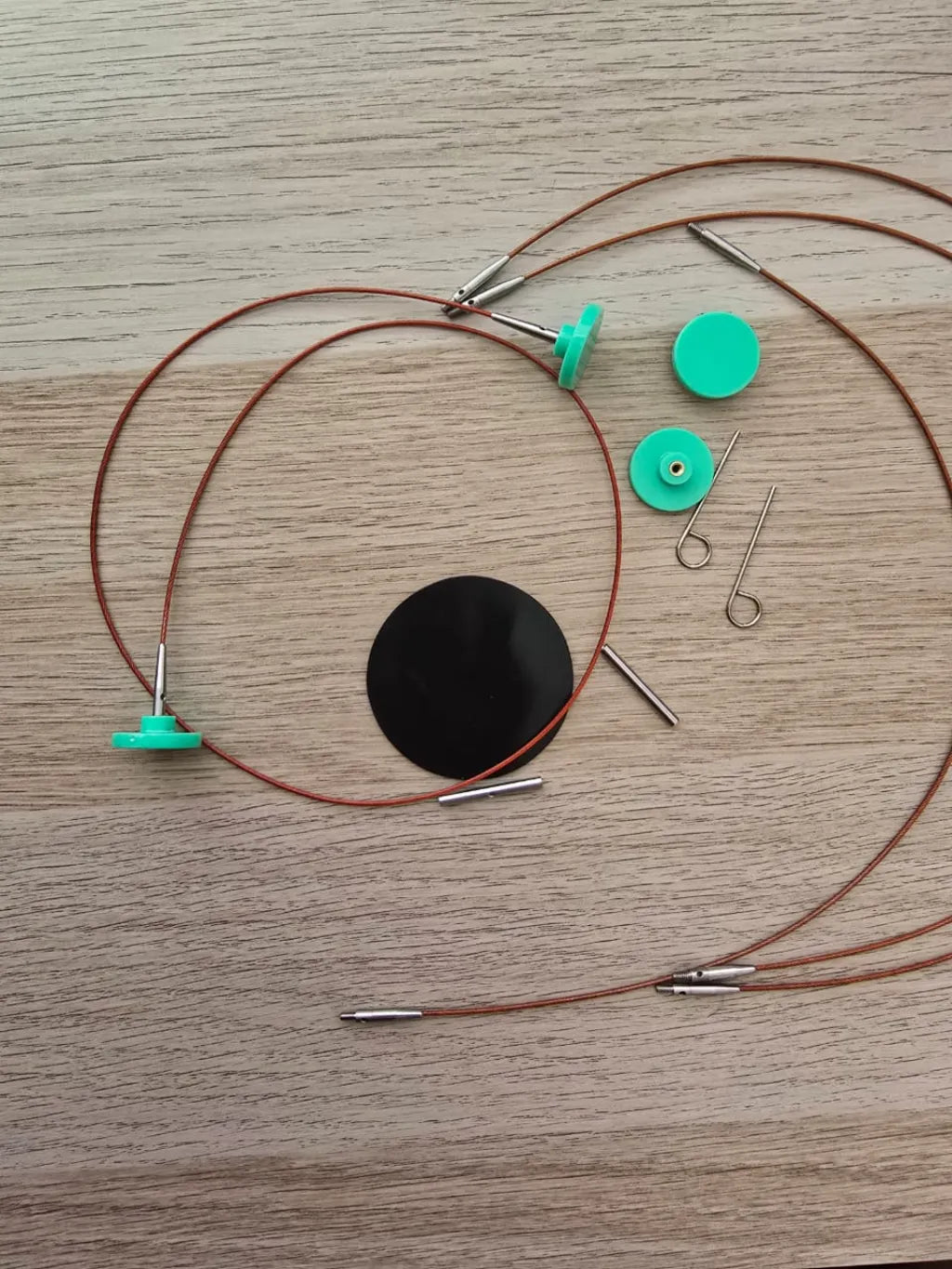 Cables de palillos circulares intercambiables color café, con topes verdes, conectores metálicos y herramientas auxiliares sobre madera.