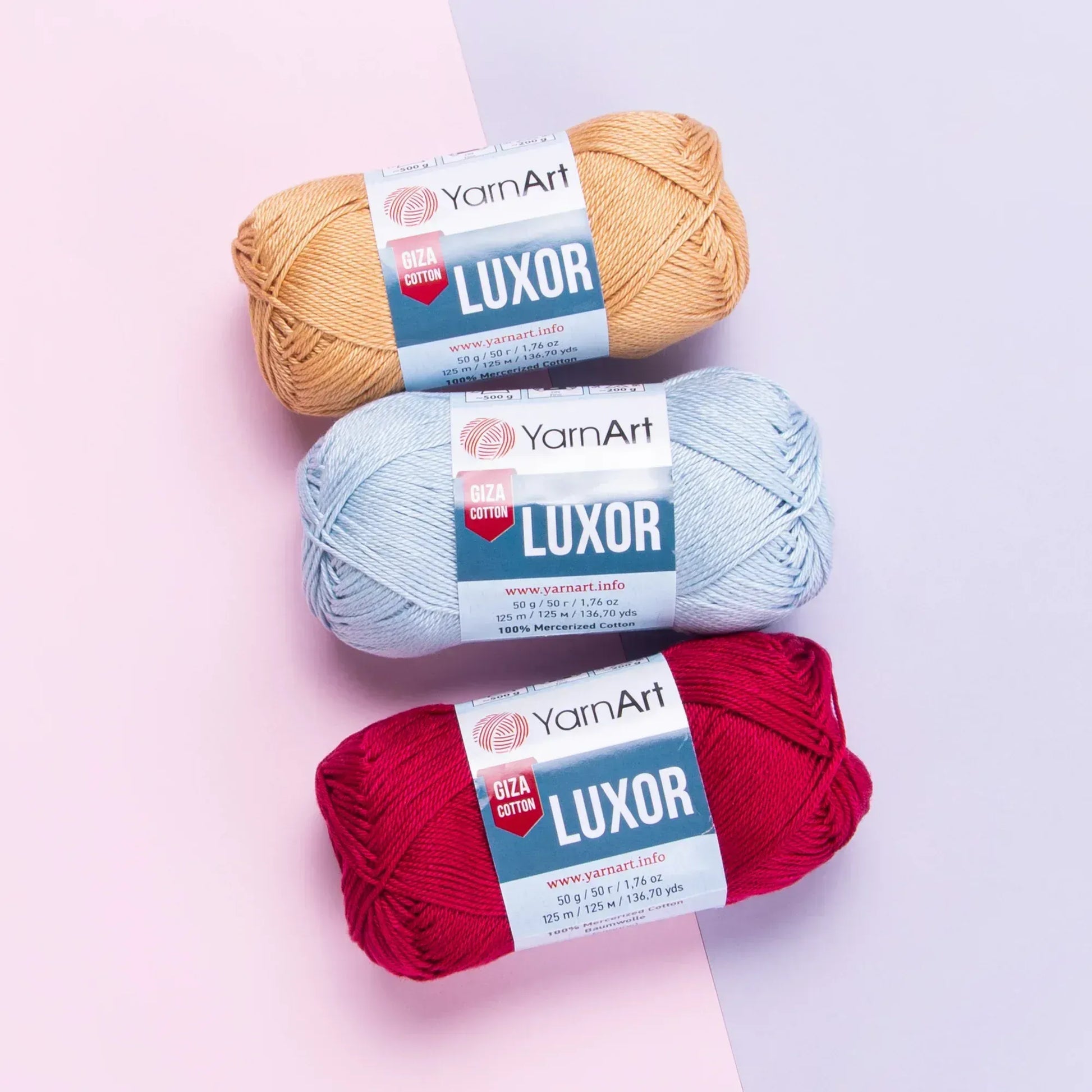 Tres ovillos de hilo YarnArt Luxor Giza Cotton de algodón mercerizado en colores beige, celeste y rojo, sobre fondo pastel.
