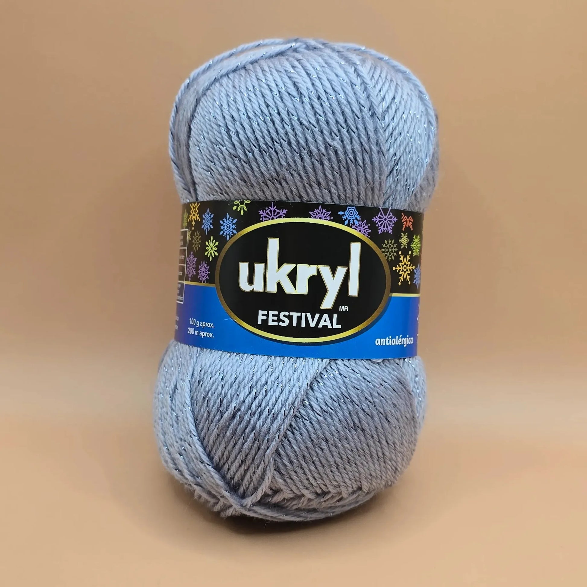 Ovillo de lana Ukryl Festival gris plateado con hilos brillantes de lurex, 94% acrílico antialérgico, fondo beige.