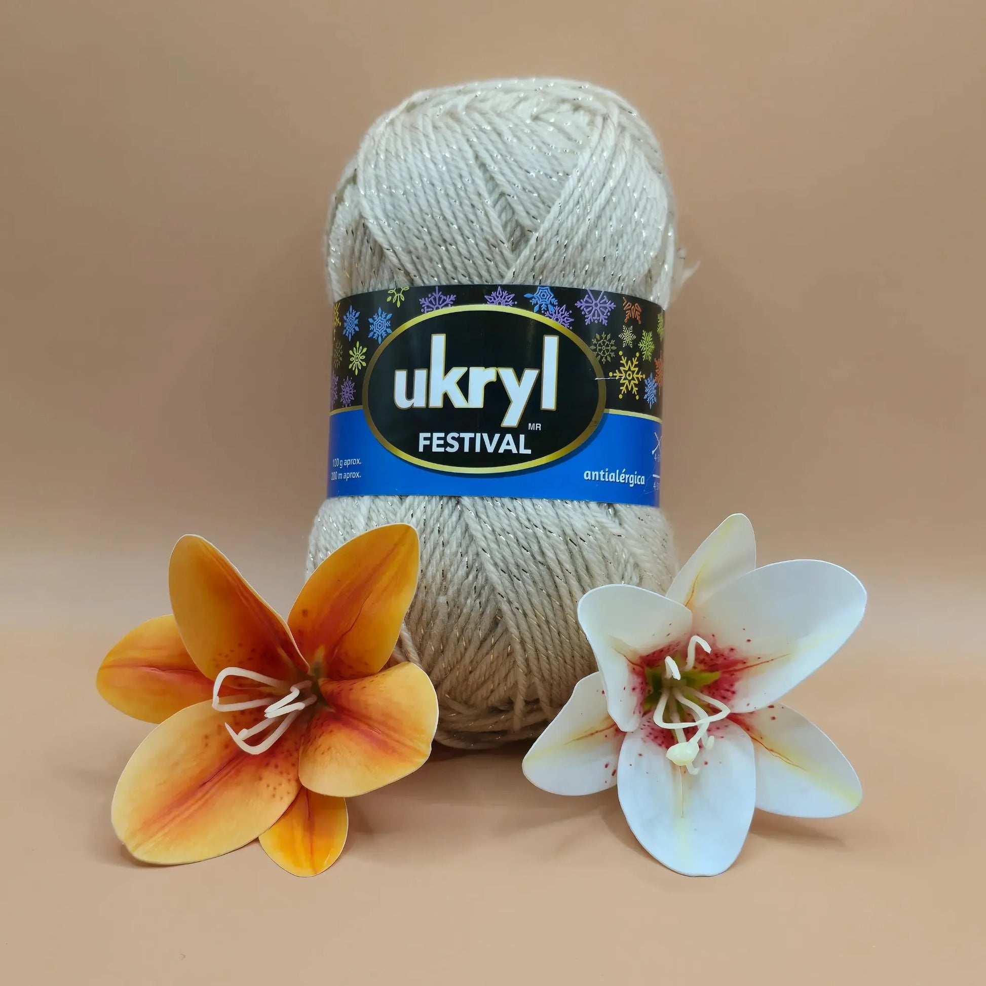 Ovillo de lana Ukryl Festival color crema con hebras de lurex plateadas, antialergica, junto a dos flores decorativas.