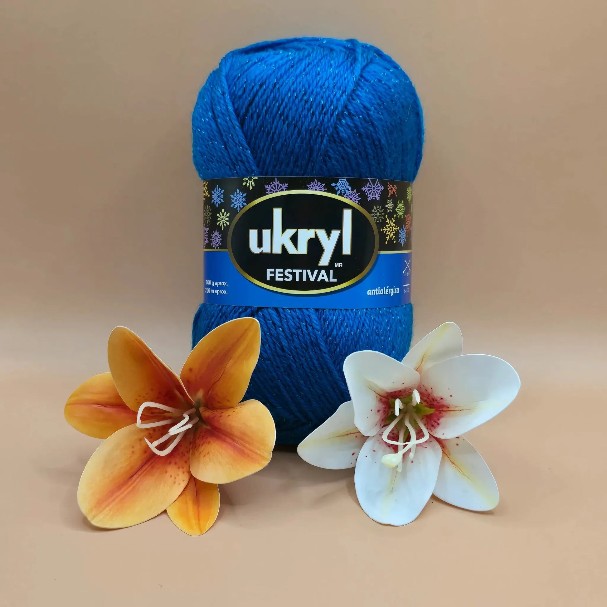 Ovillo de lana Ukryl Festival azul brillante con lúrex, 94% acrílico antialérgico, junto a flores decorativas.