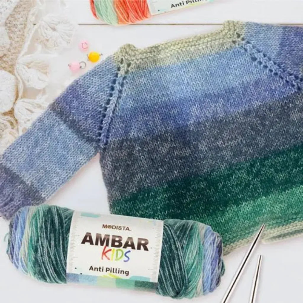 Suéter de bebé tejido a mano con lana Modista Ambar Kids de acrílico suave, colores degradados azul y verde, junto a ovillos.