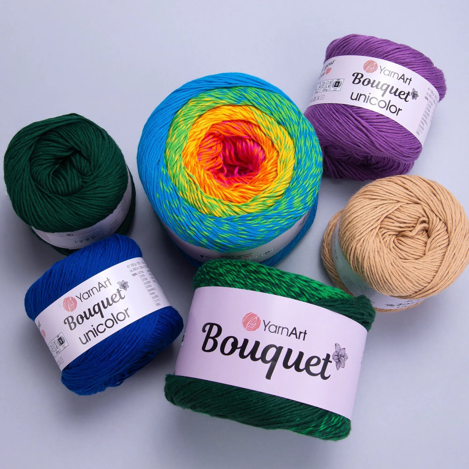 Seis ovillos de hilo YarnArt Bouquet: uno XL degradado arcoíris y cinco unicolores de algodón (verde, azul, morado, beige).