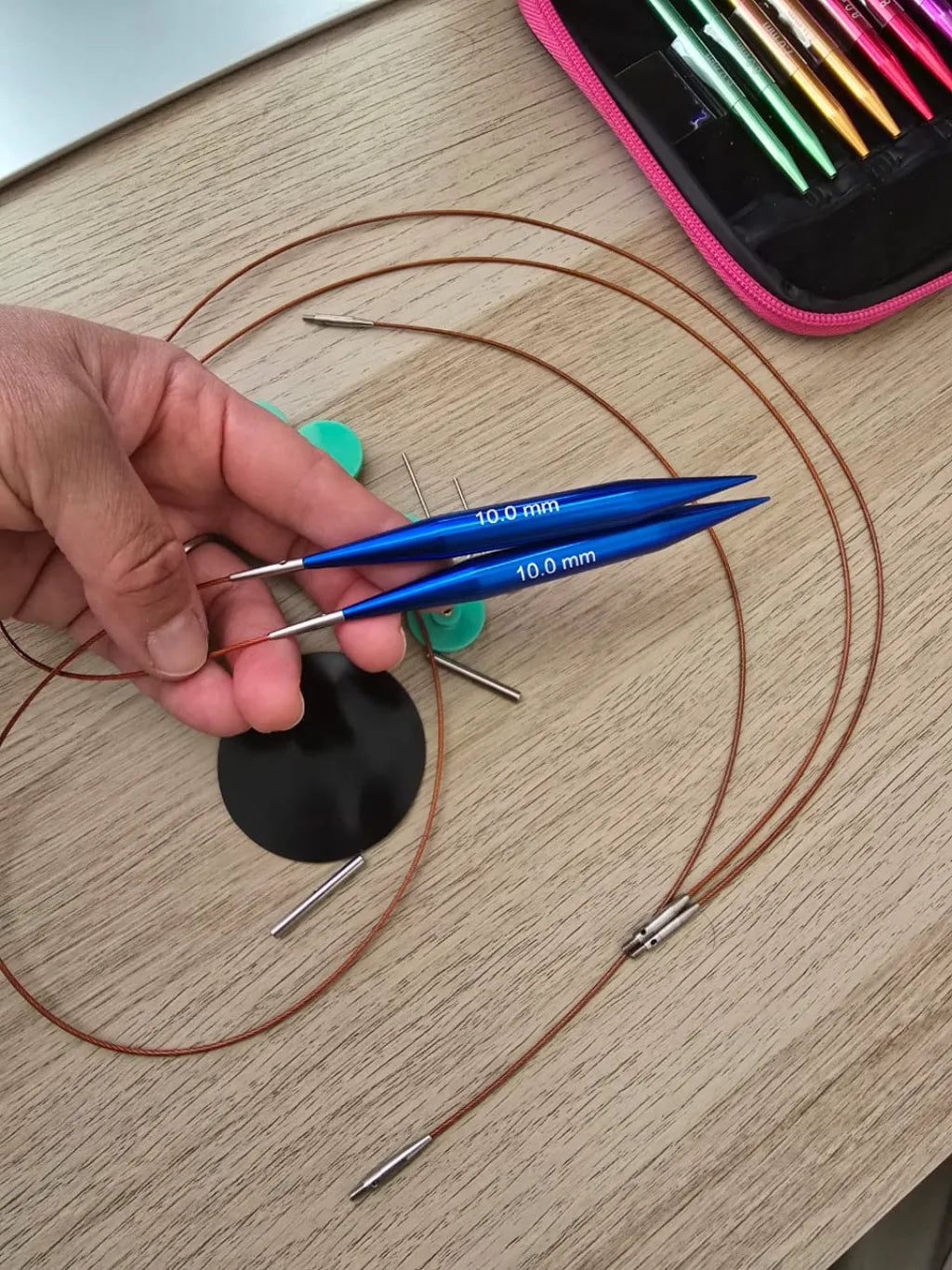 Mano sosteniendo palillos circulares intercambiables azules de aluminio de 10.0 mm con cables y accesorios sobre madera.