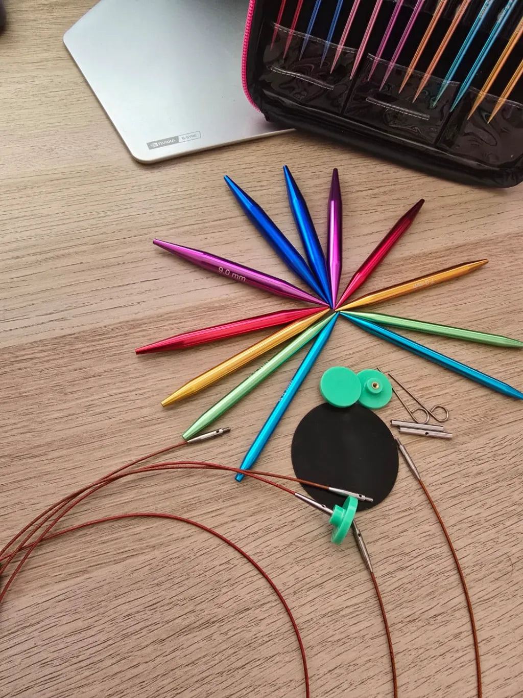 Set de palillos circulares intercambiables de aluminio en diversos colores, con cables y accesorios de tejido, sobre madera.