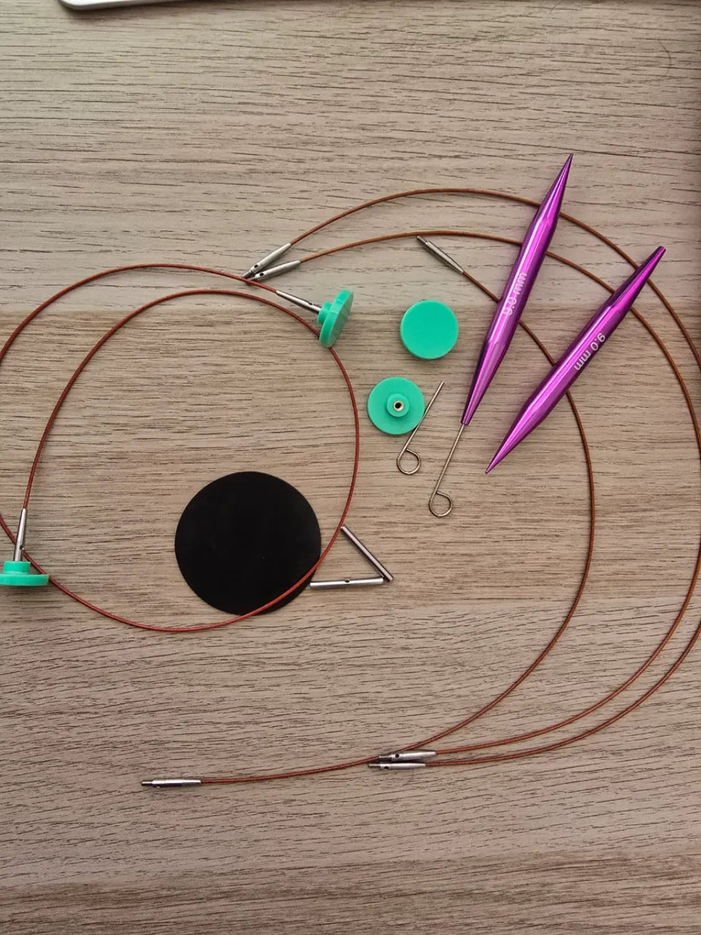 Set de palillos de tejer circulares intercambiables de aluminio color morado con cables y accesorios en una superficie de madera clara.