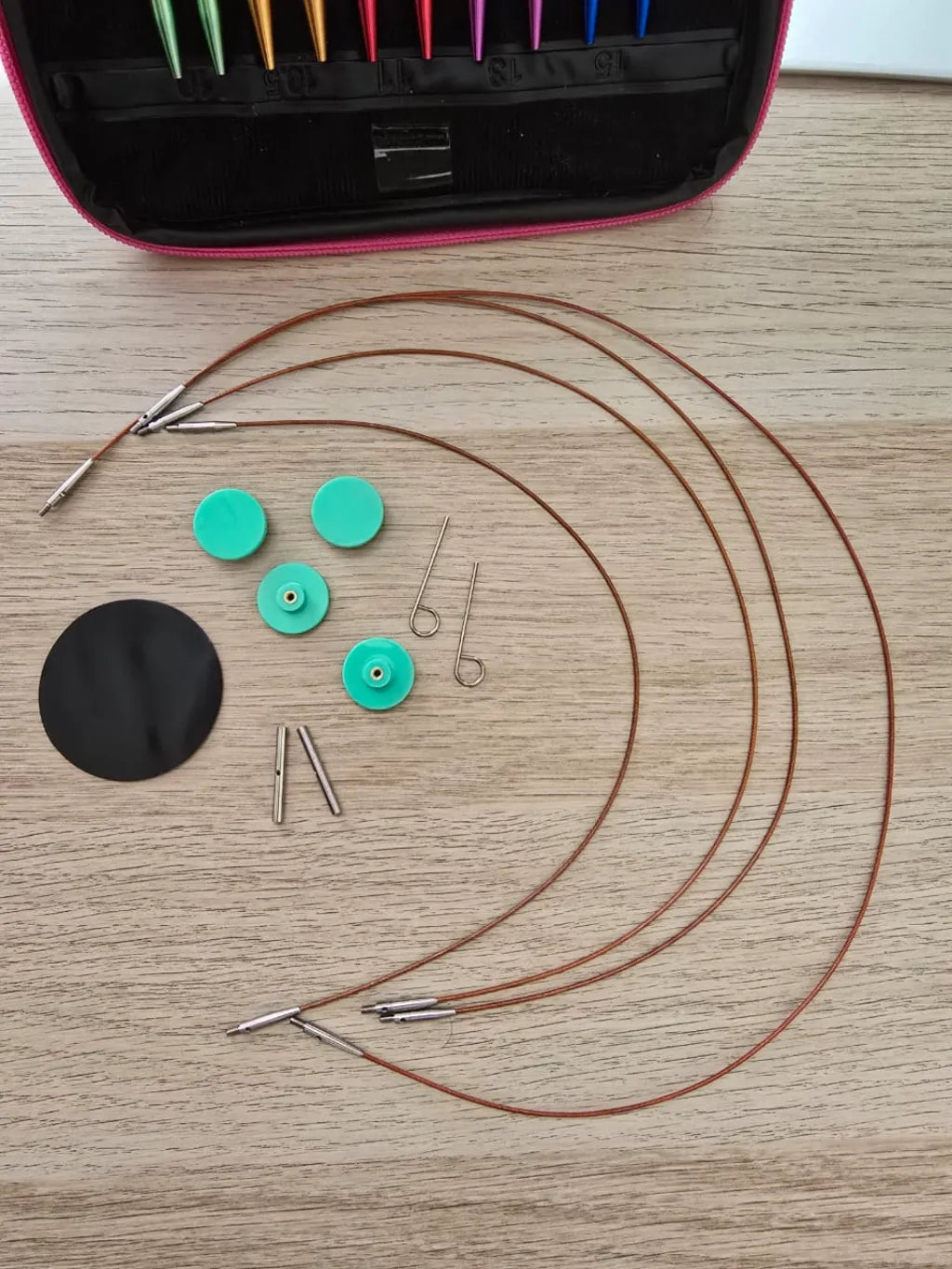 Set de palillos circulares intercambiables de aluminio con cables, topes verdes y estuche rosa sobre madera clara.