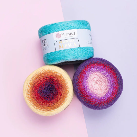 Tres ovillos de lana YarnArt Flowers Alpaca: uno turquesa y dos con degradado multicolor (naranja, morado, rosado) sobre fondo pastel.